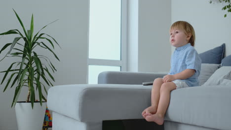 Ein-2-jähriger-Junge-Sitzt-Auf-Einem-Sofa-Und-Schaut-Fern,-Während-Er-Eine-Fernbedienung-In-Der-Hand-Hält.